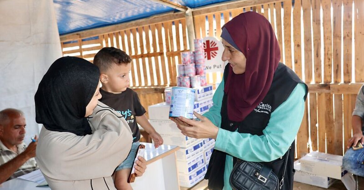 Eine Mitarbeiterin der Caritas Jerusalem zeigt einer Frau mit kleinem Kind eine Dose Babynahrung. Im Hintergrund sind Hilfspakete und das Caritas-Logo zu sehen.