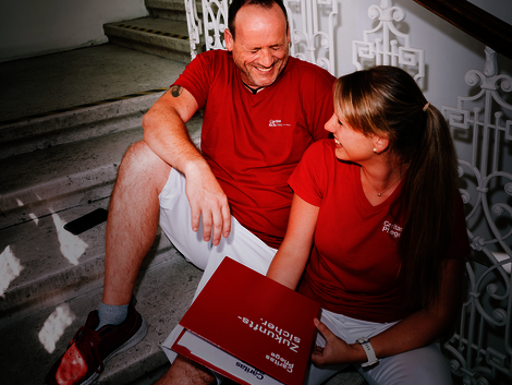 Ein Mann und eine Frau mit roten T-Shirts mit Caritas-Logo sitzen auf den Stiegen und lachen gemeinsam. Die Frau hat einen Aktenordner in der Hand.